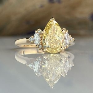 Pear-shaped yellow diamond engagement ring with two side diamonds on a gold band and a reflective surface below.