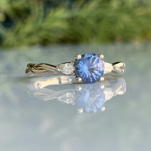Engagement ring with a round blue gemstone flanked by two small diamonds on a gold band, reflected on a mirrored surface.