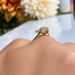 Close-up of a gold engagement ring with a diamond solitaire on a finger, against a soft floral background.