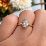 Gold engagement ring with an oval-cut diamond set in a yellow gold band, worn on a finger with a soft floral background