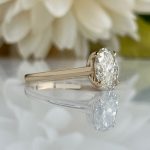 Oval-cut diamond engagement ring with a pale gold band resting on a reflective surface, background blurred with white flowers.