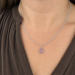 Silver necklace with a pink, haloed gemstone pendant resting at the base of the throat, worn over a brown top.