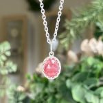 Oval pink faceted gemstone pendant on a silver chain with a beaded bezel setting, hanging against a blurred natural background.