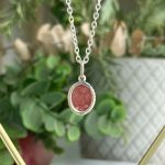 Oval red gemstone pendant on a silver chain, photographed close-up with blurred green background.