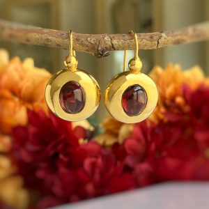 Gold-tone circular drop earrings with red cabochon stones, hanging from a branch against an autumnal blurred background.