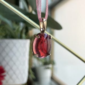 Oval pink-red gemstone pendant in a gold prong setting on a pink ribbon necklace.