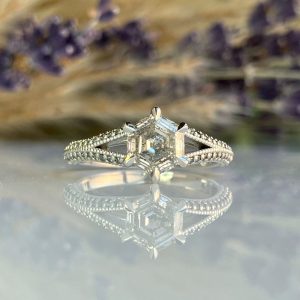 Engagement ring with a square-cut center diamond and pavé band, reflected on a glass surface.