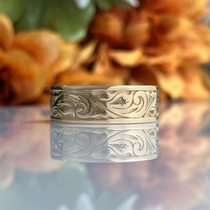 Silver engraved band ring with swirling floral motifs on a reflective surface with blurred autumn flowers in the background