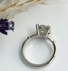 Silver engagement ring with an empty prong setting, ready for a gemstone, photographed on a light surface with dried flowers nearby.