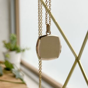 Gold-toned square pendant with rounded edges on a fine gold chain, hanging near a window.