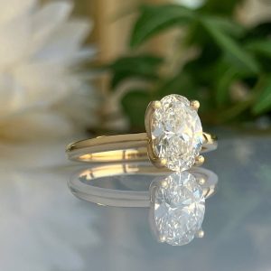 Oval-cut diamond engagement ring in a gold band, set on a reflective surface with a blurred green background.