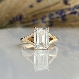 Gold ring with an emerald-cut diamond in a four-prong setting on a reflective surface.