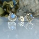 Silver stud earrings with square-cut blue gemstones in an ornate silver halo, reflected on a glossy surface.