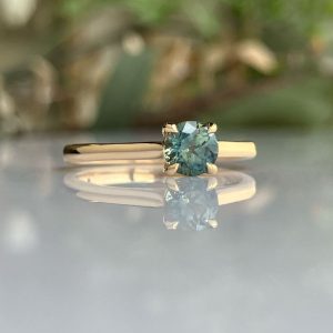 Gold ring with a square teal-blue gemstone held by four prongs, reflected on a glossy surface.