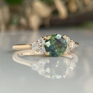 Gold ring featuring a large oval green center stone with diamonds on each side, resting on a reflective surface.