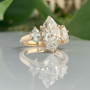 Gold ring with a large marquise-cut diamond center flanked by two smaller diamonds, reflected on a glossy surface.