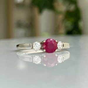 Silver ring with a red ruby center flanked by two diamonds on a reflective surface.