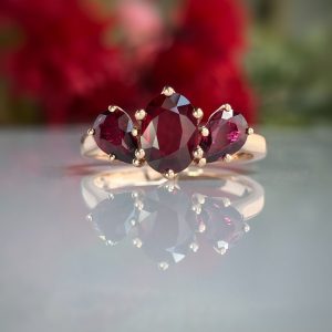 Gold ring with three red gemstones (center oval, two smaller) on a reflective surface with blurred background.