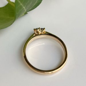 Gold ring with a small diamond solitaire in a prong setting, resting on a white surface with a green leaf in the corner