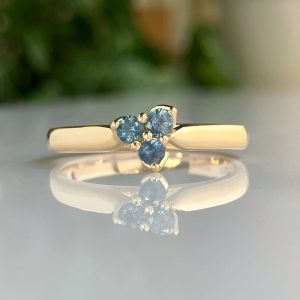 Gold ring with a trio of blue gemstones in a triangular cluster, reflecting on a glossy surface