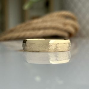 Gold wedding band with brushed center resting on a reflective surface, soft blurred background.
