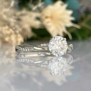 Engagement ring with a large round diamond on a twisted pavé band, reflected on a glossy surface with soft background blooms.