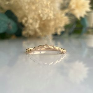 Close-up of a thin twisted rope ring in rose gold on a reflective surface with soft, blurred flowers in the background.