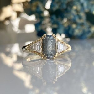 Gold ring with a rectangular blue center stone flanked by baguette-cut diamonds on a reflective surface.