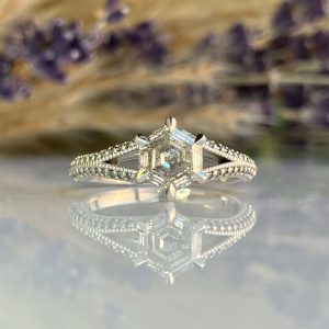 Engagement ring with a square-cut center diamond and pavé band, reflected on a mirror-like surface.