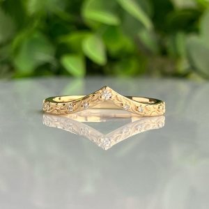 Gold ring with engraved leaf pattern and small diamonds along the band, placed on a reflective surface with green foliage in the background.