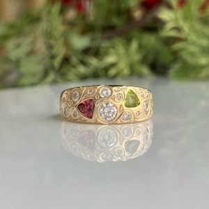 Gold ring with multiple gemstones (diamonds and colored stones) resting on a shiny reflective surface; blurred green background behind.