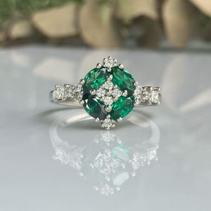 Close-up of a silver ring with a large green emerald surrounded by a halo of diamonds, on a reflective surface with a blurred background.