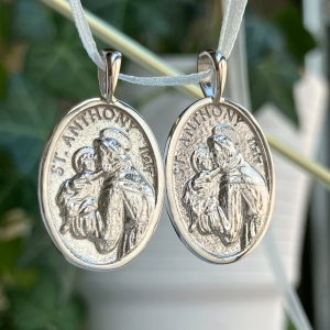 Two identical oval silver St. Anthony medals hanging side by side on light cords, each showing Saint Anthony cradling the Christ child.