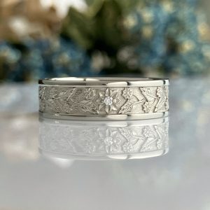 Silver ring with an engraved floral pattern and a small central gemstone, resting on a reflective surface.
