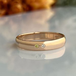 Gold band ring with two small stones (lime-green and clear) set side by side on a polished surface, reflecting below.