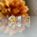 Silver stud earrings with a yellow gemstone set in a prong setting, reflected on a glossy surface with a warm blurred background.
