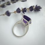 Silver ring with a large purple rectangular amethyst gemstone, set in a prong setting on a light surface with lavender sprigs nearby.