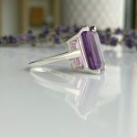 Buttress-cut purple amethyst ring set in silver with a sleek band and a reflective surface nearby