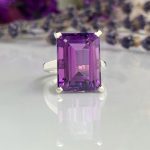 Silver ring featuring a large square-cut purple amethyst, shown on a reflective surface in a close-up shot.
