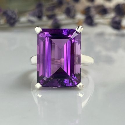 Emerald-cut purple amethyst ring in a four-prong silver setting on a reflective surface.